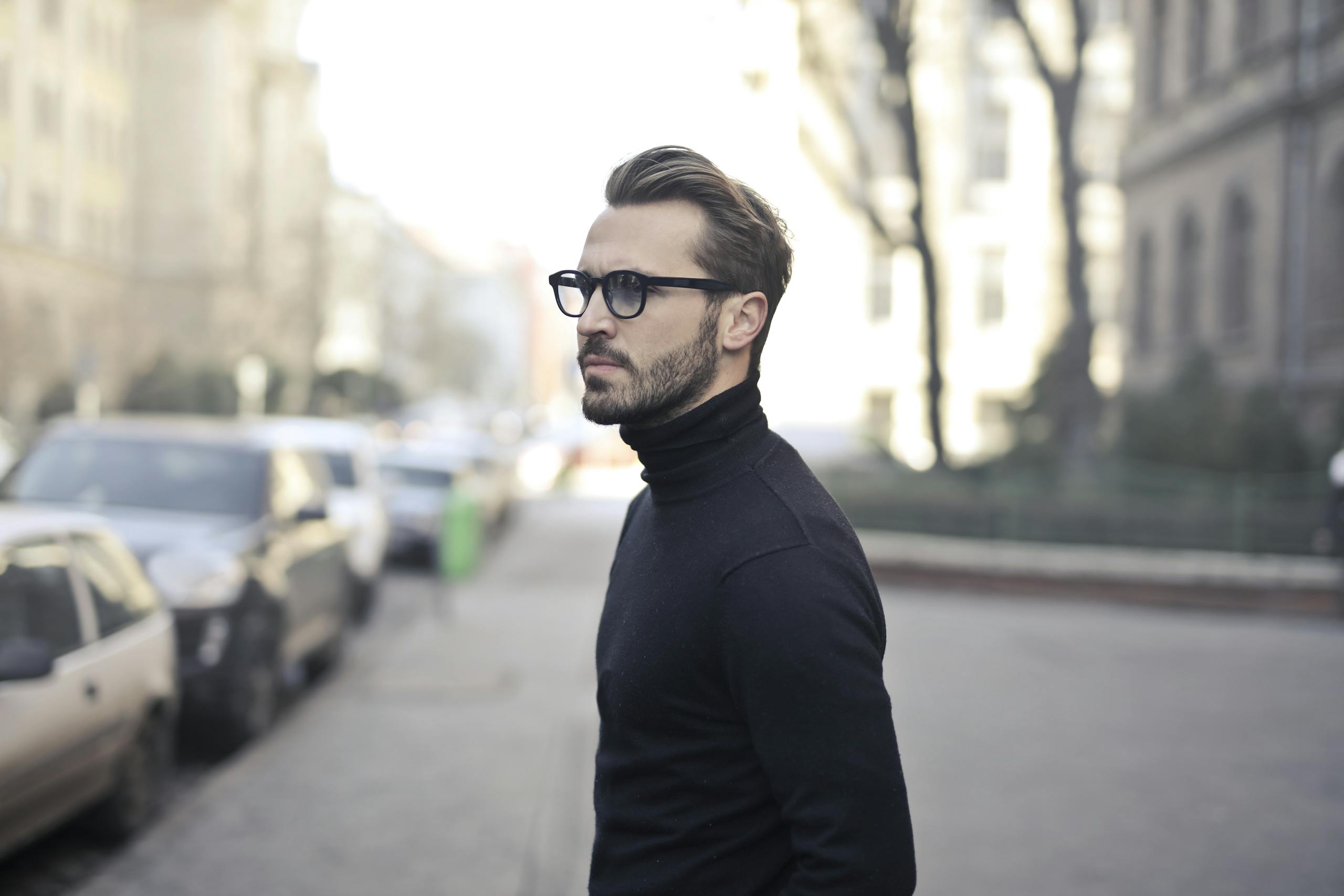 A fashionable man in a black top and glasses poses in an urban street with parked cars.